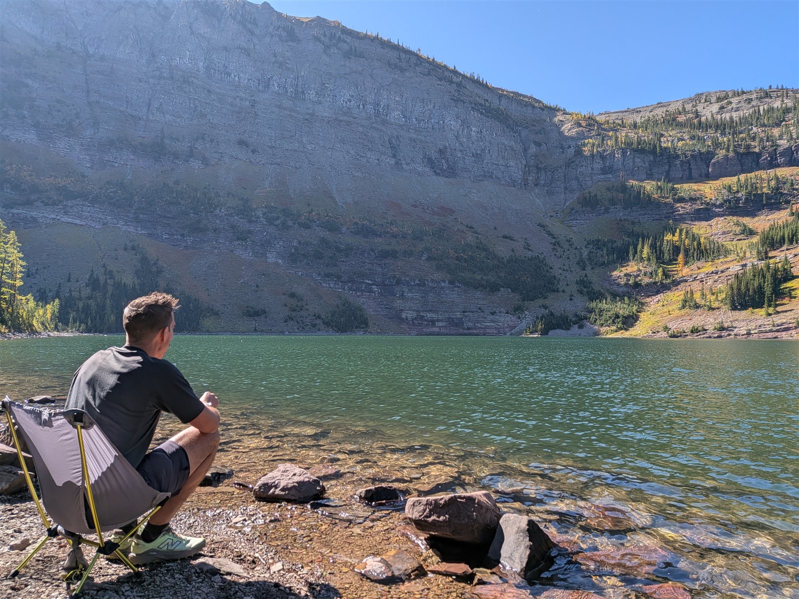 A backpacker relaxing in a chair beside Lower Twin Lake beneath a tall cliff