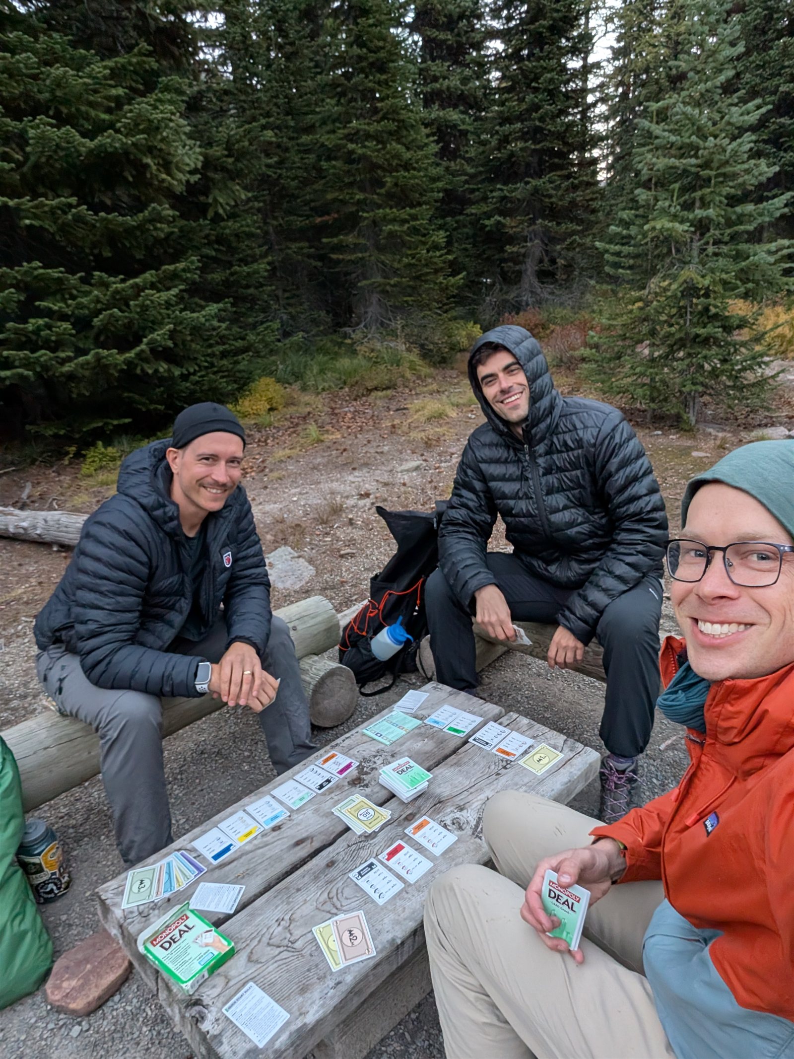 Three backpackers playing Monopoly Deal around a wooden table at a forest campsite