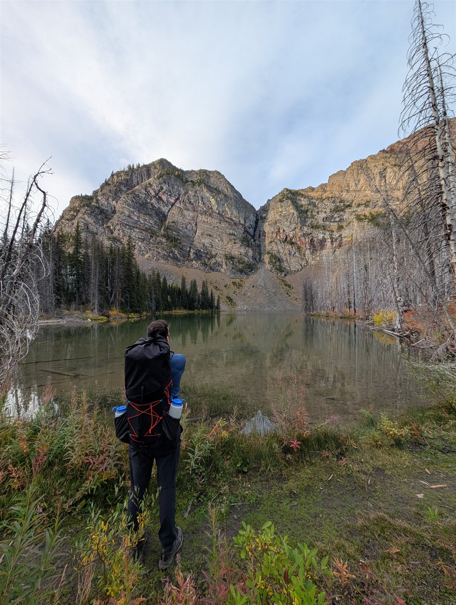 A backpacker standing beside still Goat Lake beneath steep rocky cliffs and a pale sky
